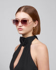 In a plain grey setting, a short-haired woman exudes sophistication in For Art's Sake® Dusk pink-tinted, chunky cat-eye sunglasses and a black halter dress with a floral accent, gazing to the side.