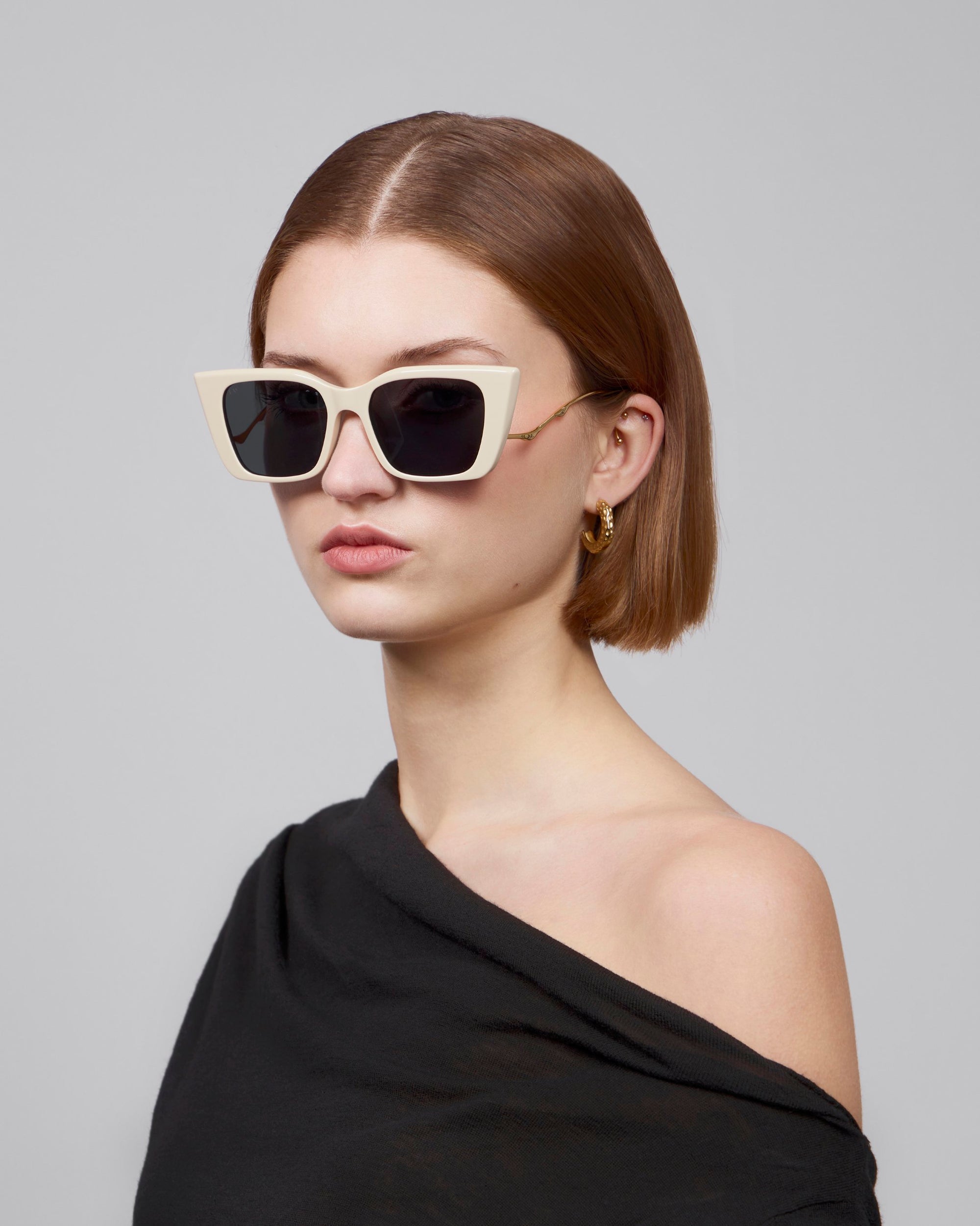 A person with short brown hair wears For Art's Sake® Dusk large white cat-eye sunglasses with an acetate front. They pair them with gold hoop earrings and have a serious expression, dressed in a black off-the-shoulder top against a gray background.