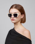 A person with short brown hair wears For Art's Sake® Dusk large white cat-eye sunglasses with an acetate front. They pair them with gold hoop earrings and have a serious expression, dressed in a black off-the-shoulder top against a gray background.