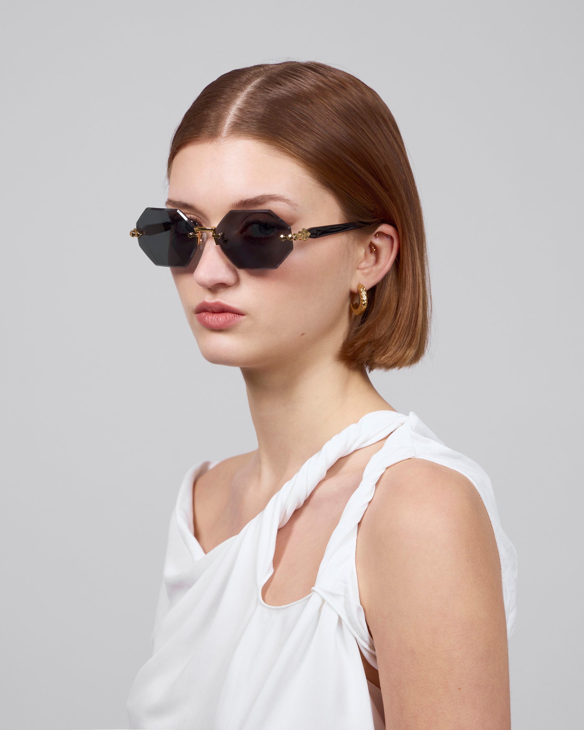 A person with short brown hair wears Haze cat-eye sunglasses by For Art's Sake® and gold hoop earrings from the Rose Garden collection, dressed in a white asymmetrical top against a plain gray background.