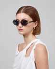 A person with short brown hair wears Haze cat-eye sunglasses by For Art's Sake® and gold hoop earrings from the Rose Garden collection, dressed in a white asymmetrical top against a plain gray background.