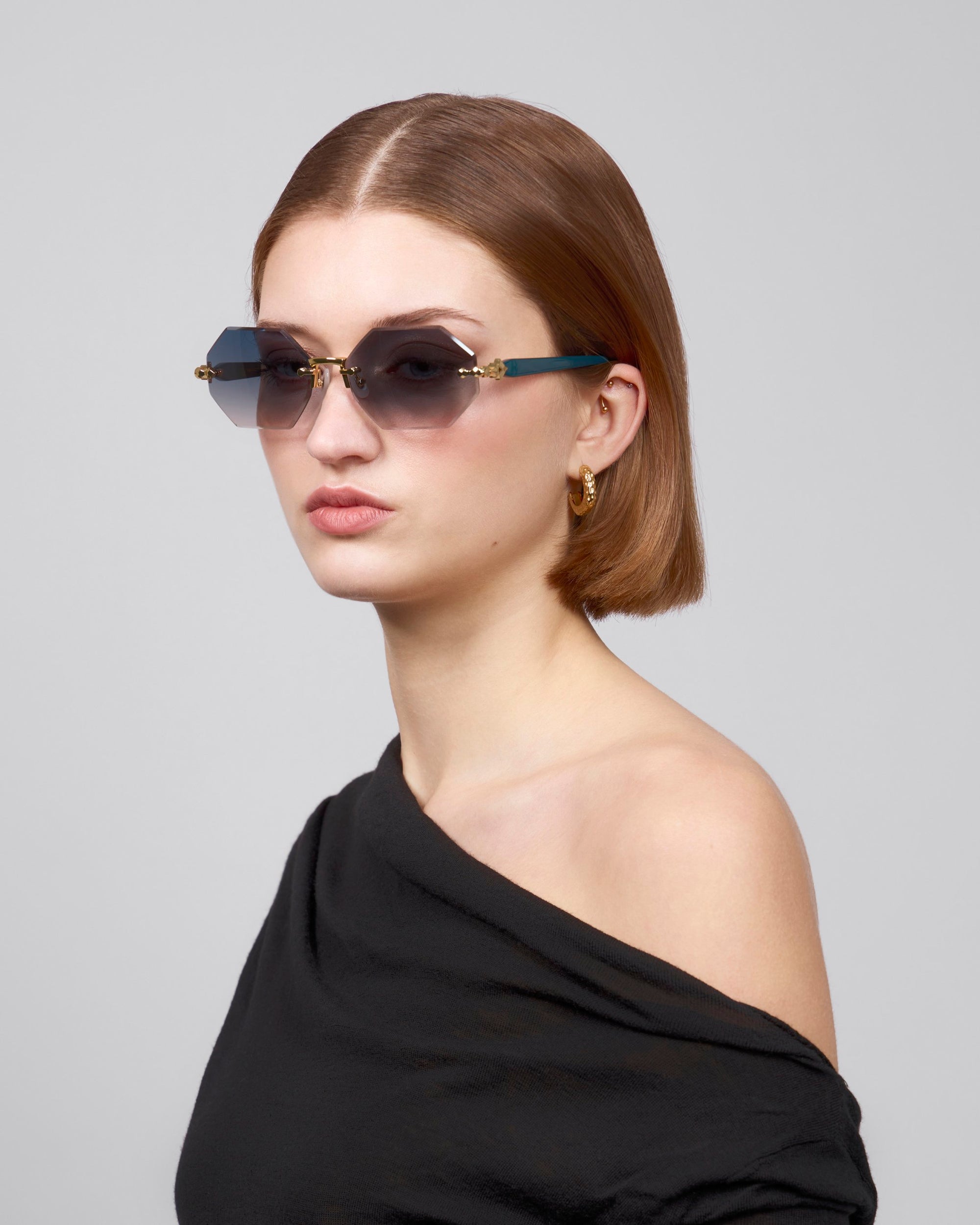 A woman with short brown hair and rimless lenses poses stylishly against a gray background, wearing gold hoop earrings and an asymmetrical black top from the Rose Garden collection by For Art's Sake®, named Haze.