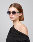 A woman with short brown hair and rimless lenses poses stylishly against a gray background, wearing gold hoop earrings and an asymmetrical black top from the Rose Garden collection by For Art's Sake®, named Haze.