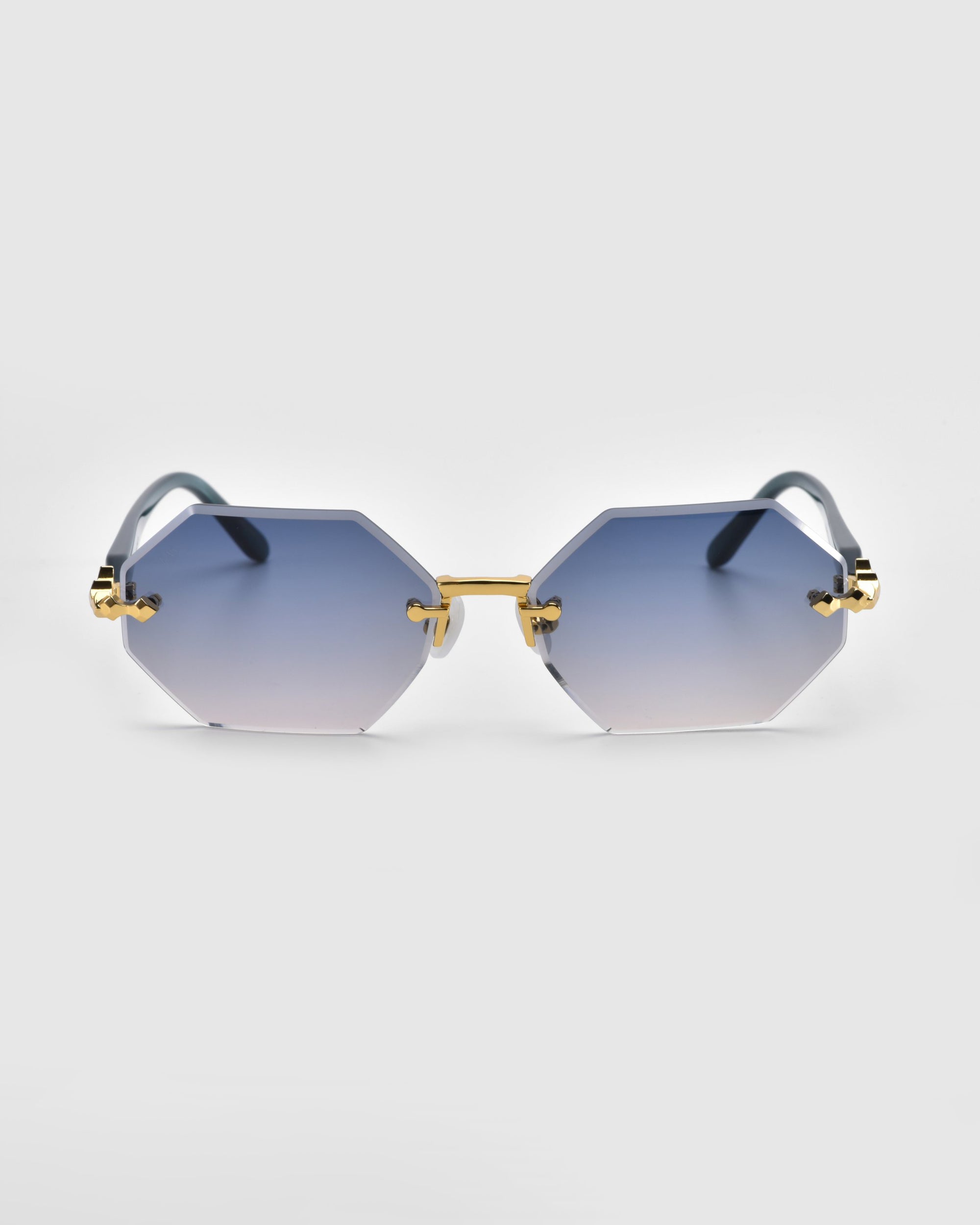 Part of For Art's Sake® Rose Garden collection, the Haze sunglasses feature a hexagonal shape with gradient blue tint, gold-tone hardware, sleek bridge, and subtle temple decorations. Their sophisticated design is showcased by rimless lenses against a plain white background.