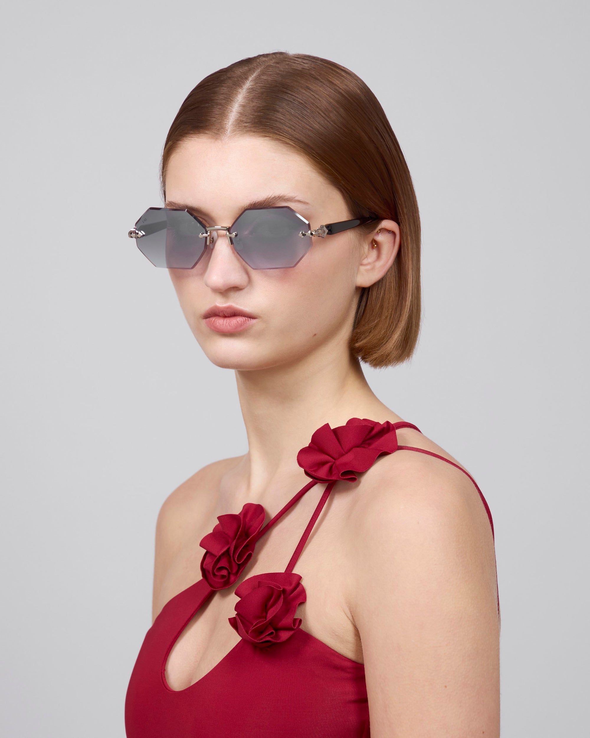 A woman with shoulder-length brown hair wears For Art's Sake® Haze geometric rimless sunglasses and a red dress with floral strap details from the Rose Garden collection, facing slightly to the side against a plain background.