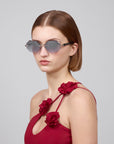 A woman with shoulder-length brown hair wears For Art's Sake® Haze geometric rimless sunglasses and a red dress with floral strap details from the Rose Garden collection, facing slightly to the side against a plain background.