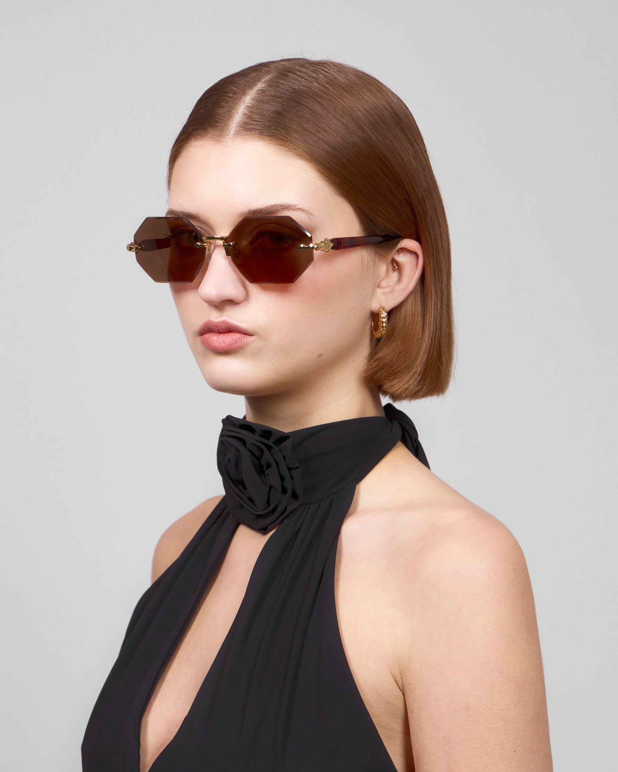 A person with short brown hair wears hexagonal Haze sunglasses from For Art's Sake® and a black sleeveless top with floral neck detail, looking sideways against a plain background.