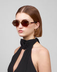 A person with short brown hair wears hexagonal Haze sunglasses from For Art's Sake® and a black sleeveless top with floral neck detail, looking sideways against a plain background.