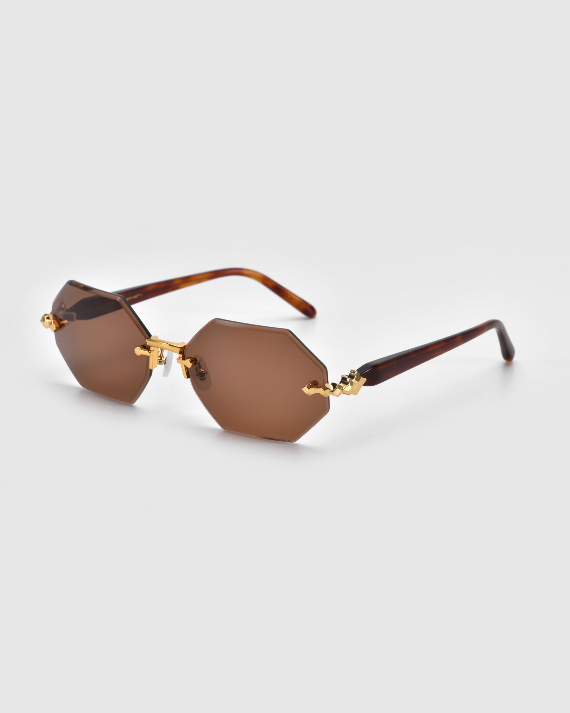 Hexagonal brown "Haze" sunglasses by For Art's Sake®, featuring gold accents and elegant temple decorations from the Rose Garden collection, displayed on a light gray background.