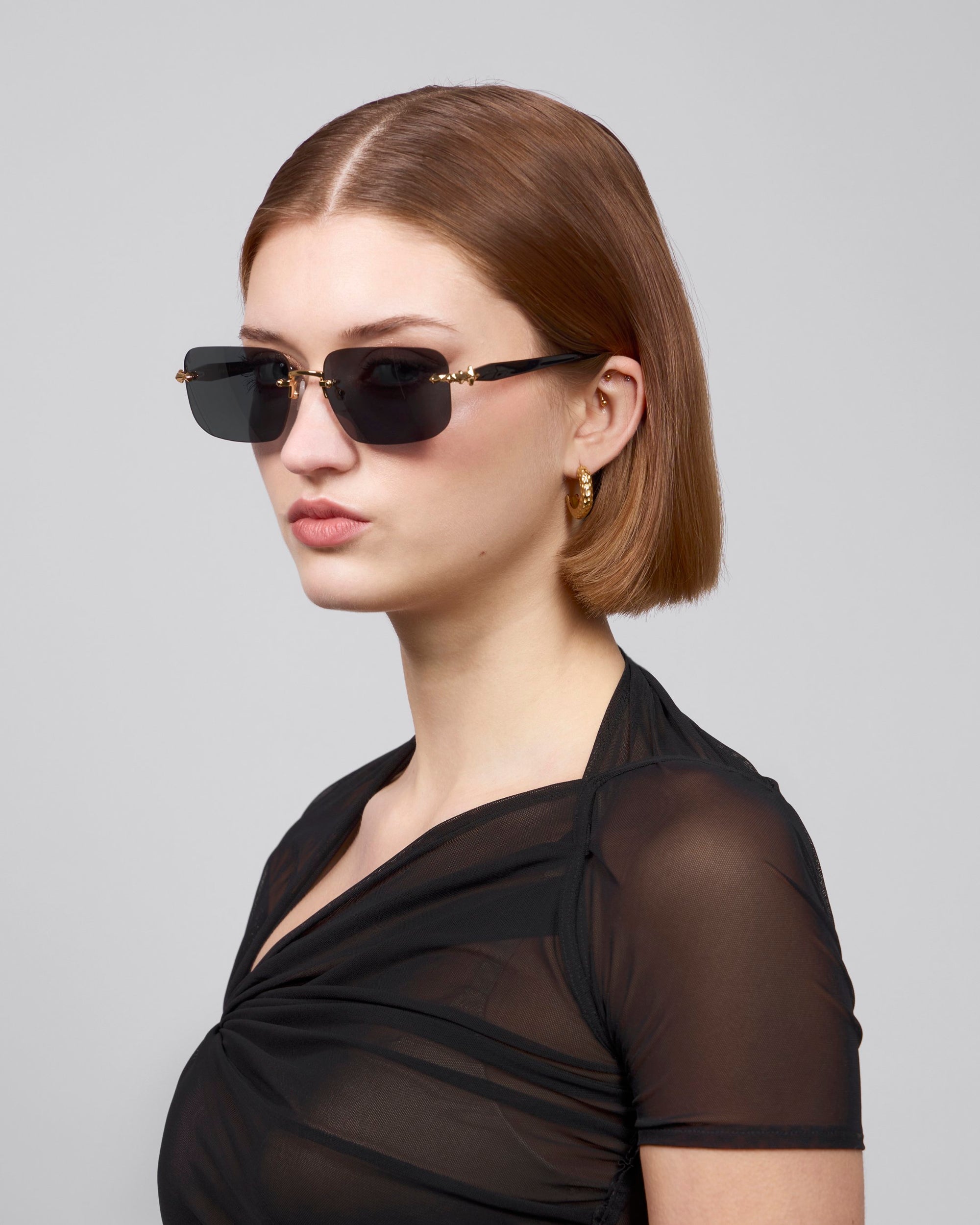 A person with short brown hair and dark rectangular lenses wears the sheer black "Edith" top from For Art's Sake® against a gray background, embodying contemporary style.