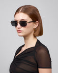 A person with short brown hair and dark rectangular lenses wears the sheer black "Edith" top from For Art's Sake® against a gray background, embodying contemporary style.