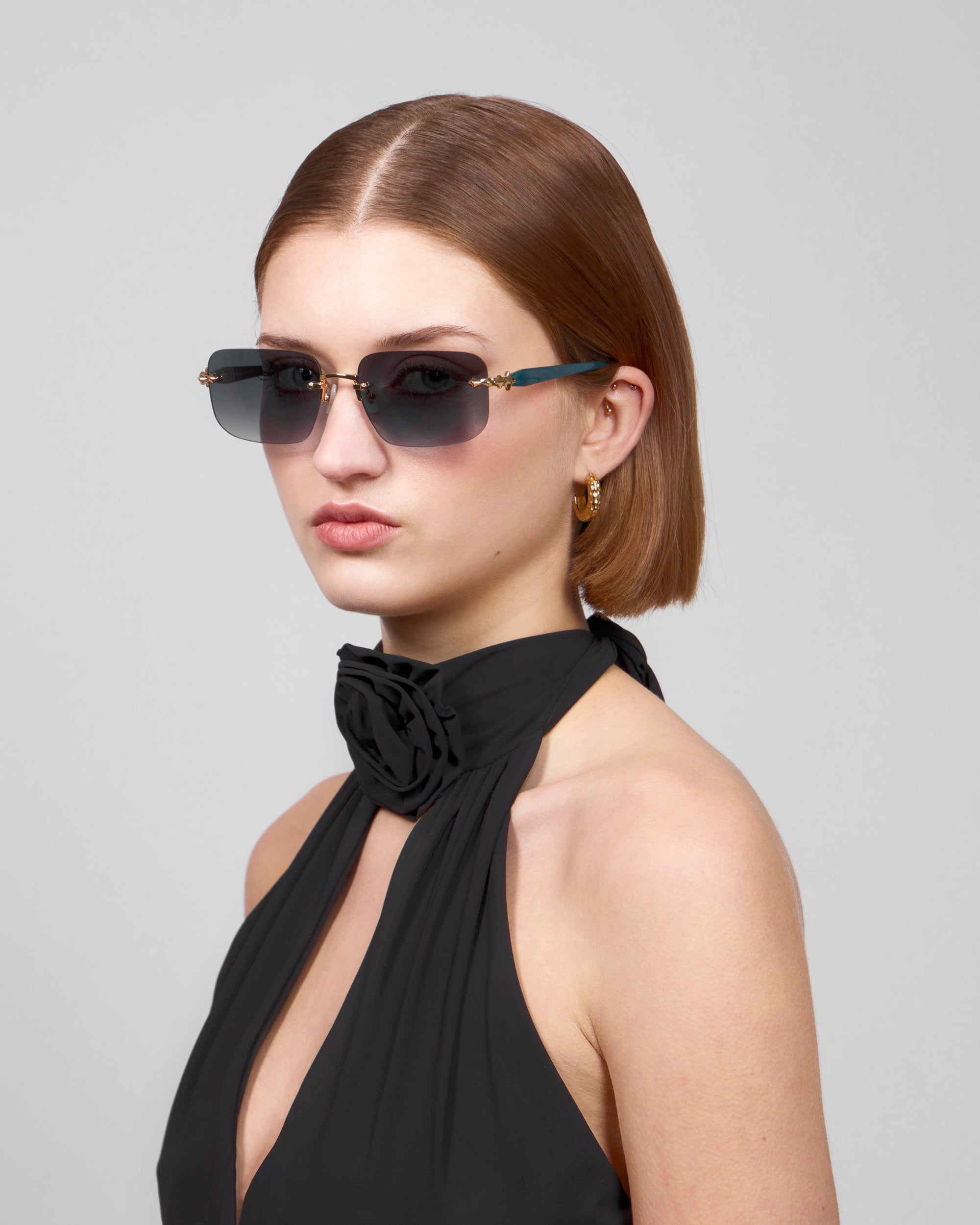 A person with short brown hair and a serious expression poses against a plain gray background, wearing chic rectangular sunglasses by For Art's Sake® and the black Edith halter dress from the Rose Garden collection, featuring a rosette at the neck. Gold hoop earrings complete their look.