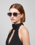 A person with short brown hair and a serious expression poses against a plain gray background, wearing chic rectangular sunglasses by For Art's Sake® and the black Edith halter dress from the Rose Garden collection, featuring a rosette at the neck. Gold hoop earrings complete their look.