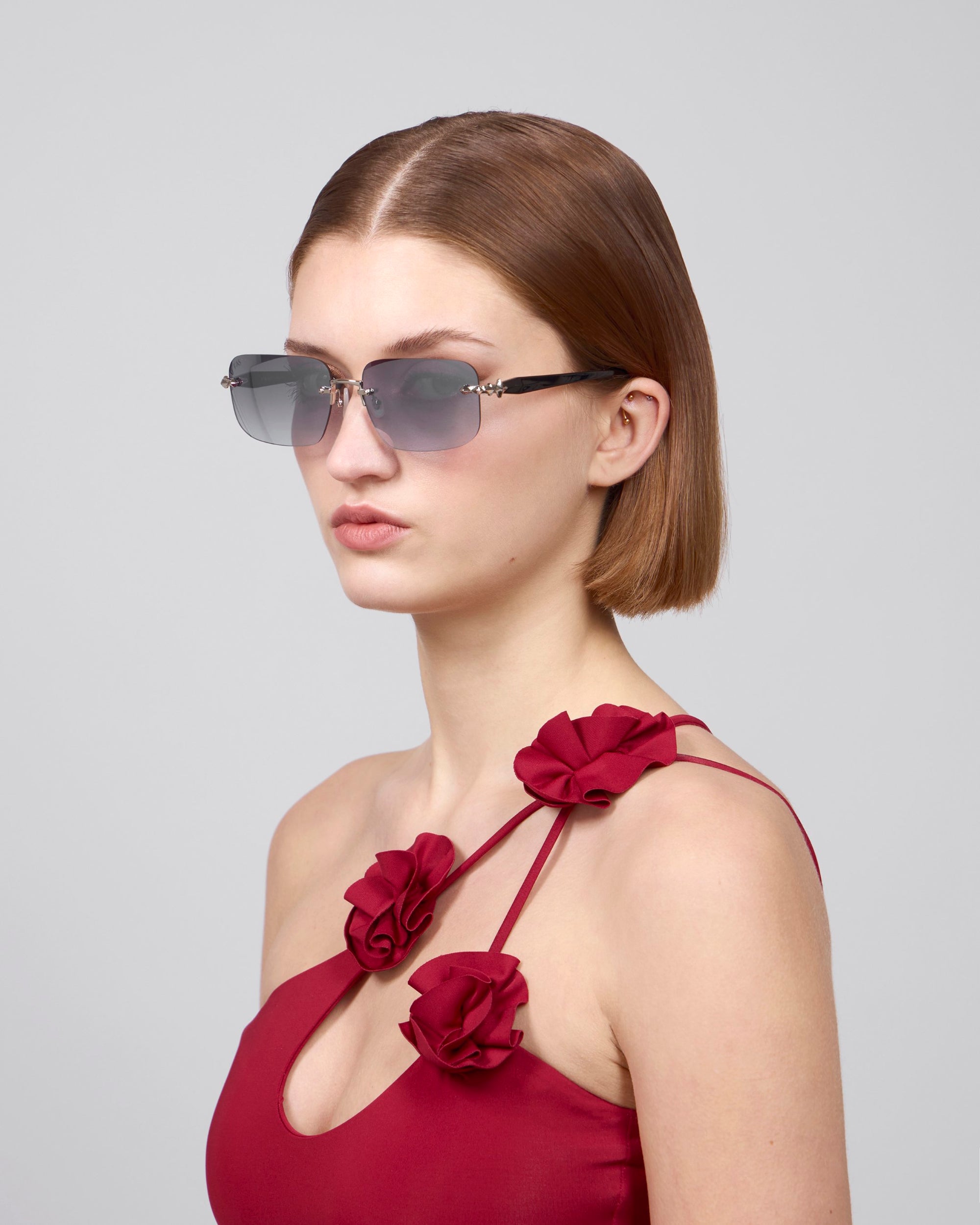 Against a plain backdrop, a woman with shoulder-length brown hair and sunglasses featuring rectangular lenses exudes contemporary style in the Edith from For Art's Sake®, wearing a red dress with rose-detailed straps, perfectly capturing the essence of the Rose Garden collection.