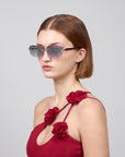 Against a plain backdrop, a woman with shoulder-length brown hair and sunglasses featuring rectangular lenses exudes contemporary style in the Edith from For Art's Sake®, wearing a red dress with rose-detailed straps, perfectly capturing the essence of the Rose Garden collection.