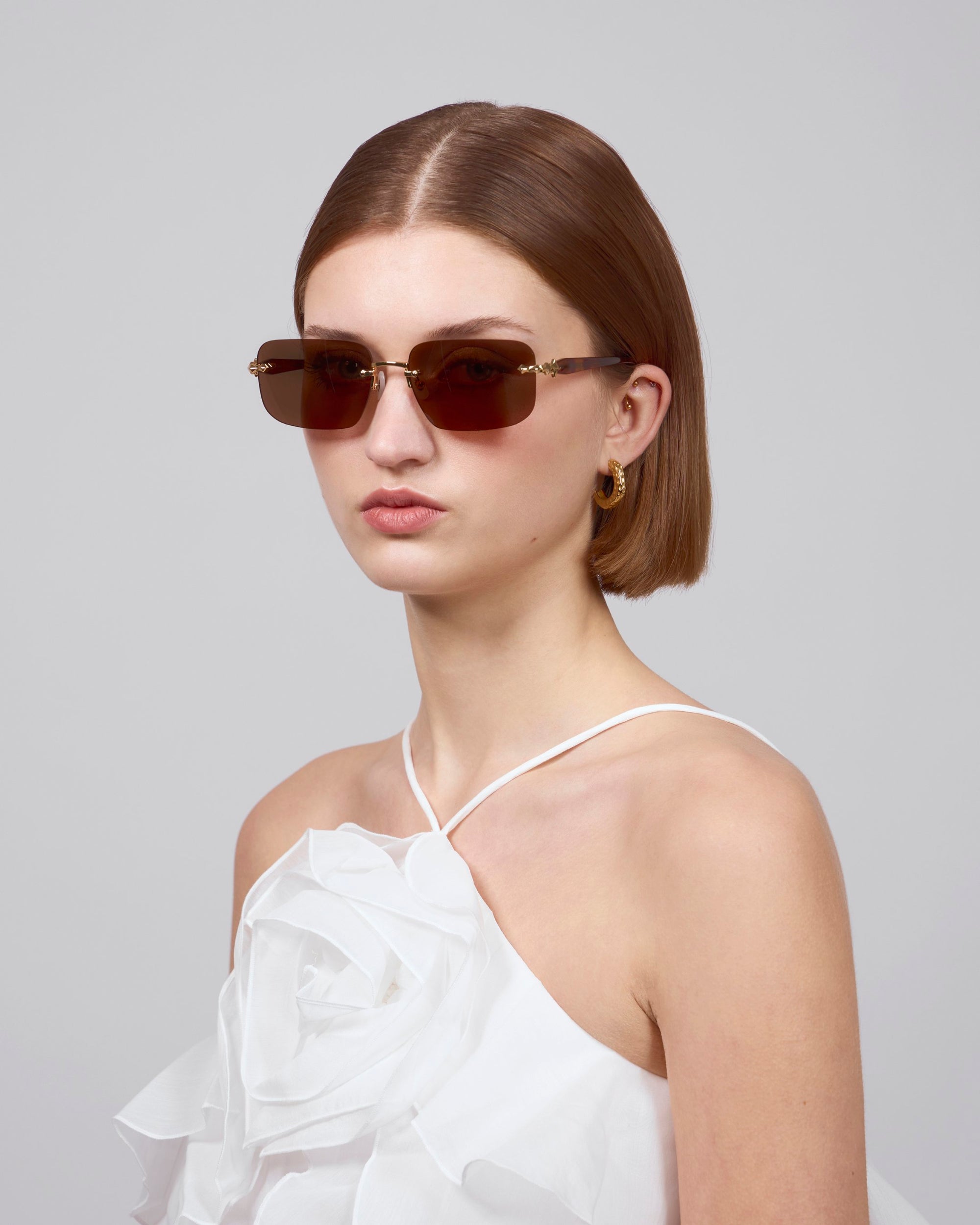 A person wears For Art's Sake® Edith rectangular-lensed sunglasses, a white halter top with large fabric flowers from the Rose Garden collection, and gold hoop earrings. Their straight, chin-length hair stands out against the plain gray background, enhancing their contemporary style.