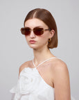 A person wears For Art's Sake® Edith rectangular-lensed sunglasses, a white halter top with large fabric flowers from the Rose Garden collection, and gold hoop earrings. Their straight, chin-length hair stands out against the plain gray background, enhancing their contemporary style.