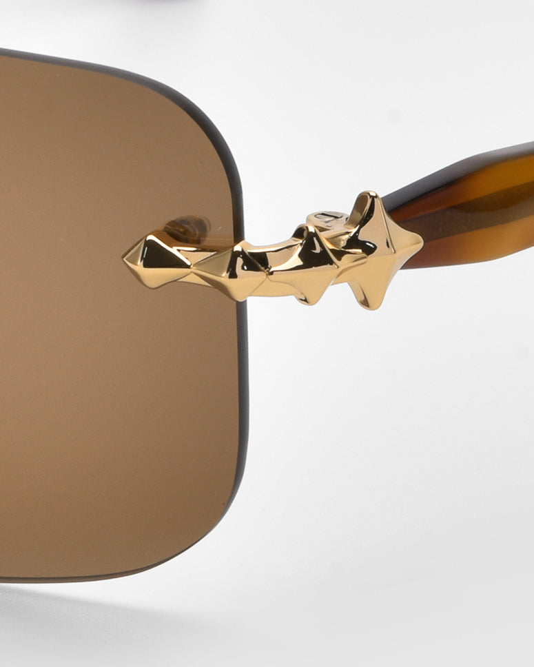 Close-up of For Art's Sake® Edith sunglasses from the Rose Garden collection, showcasing a unique gold textured star design on the arms. These brown sunglasses feature soft rectangular lenses for a timeless classic look against a gentle gray background.