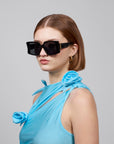 With shoulder-length brown hair, the person sports oversized black acetate sunglasses by For Art's Sake®, paired with a sleeveless blue top featuring floral shoulder details. The plain gray backdrop highlights their chic Bloom ensemble.