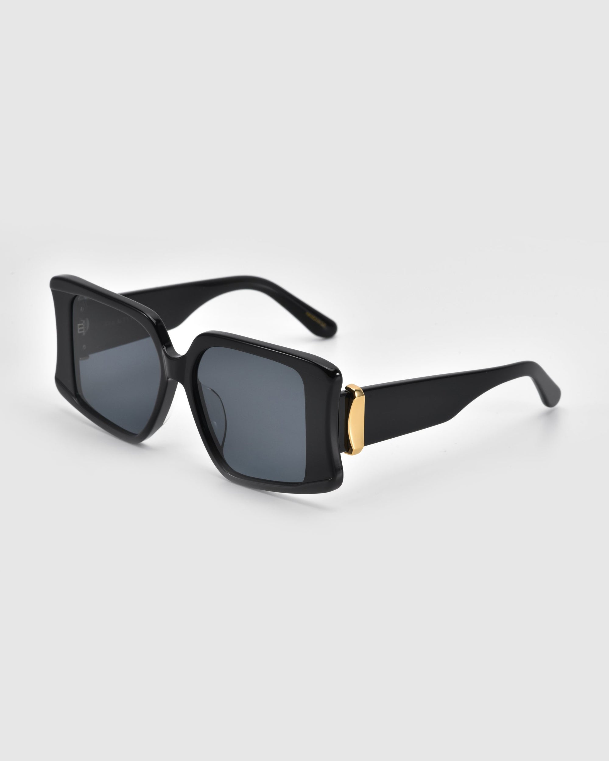 The Bloom sunglasses by For Art's Sake® are black rectangular frames with dark lenses and gold accents on the temples, resembling jewelry-like metal rings, displayed against a light gray background.