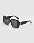 The Bloom sunglasses by For Art's Sake® are black rectangular frames with dark lenses and gold accents on the temples, resembling jewelry-like metal rings, displayed against a light gray background.