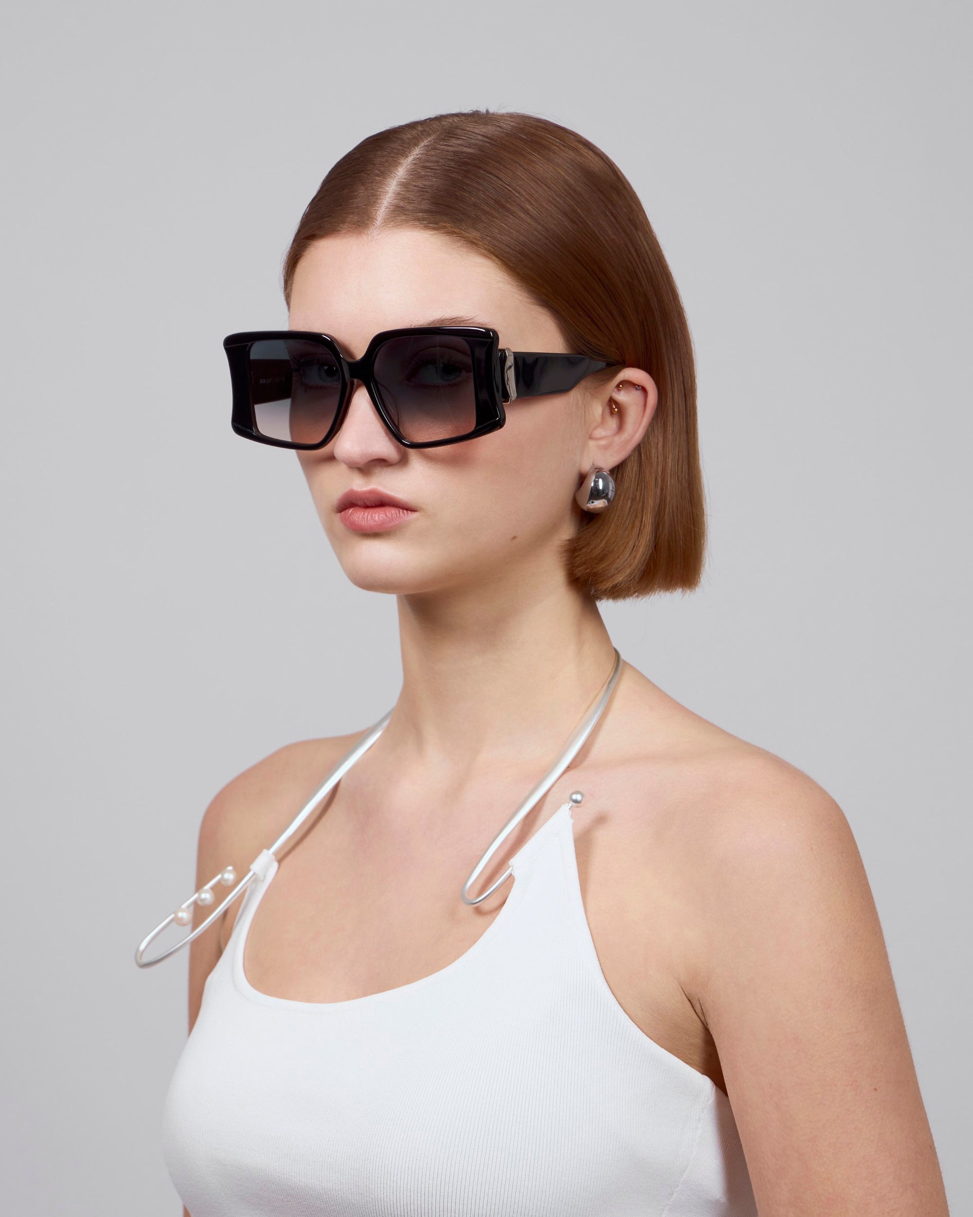 A person with short brown hair wears oversized Bloom acetate glasses by For Art's Sake® and pearl earrings, paired with a white spaghetti strap top accented by a silver chain. The plain gray background highlights the bold statement eyewear's premium craftsmanship.