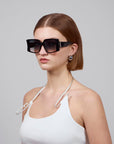 A person with short brown hair wears oversized Bloom acetate glasses by For Art's Sake® and pearl earrings, paired with a white spaghetti strap top accented by a silver chain. The plain gray background highlights the bold statement eyewear's premium craftsmanship.