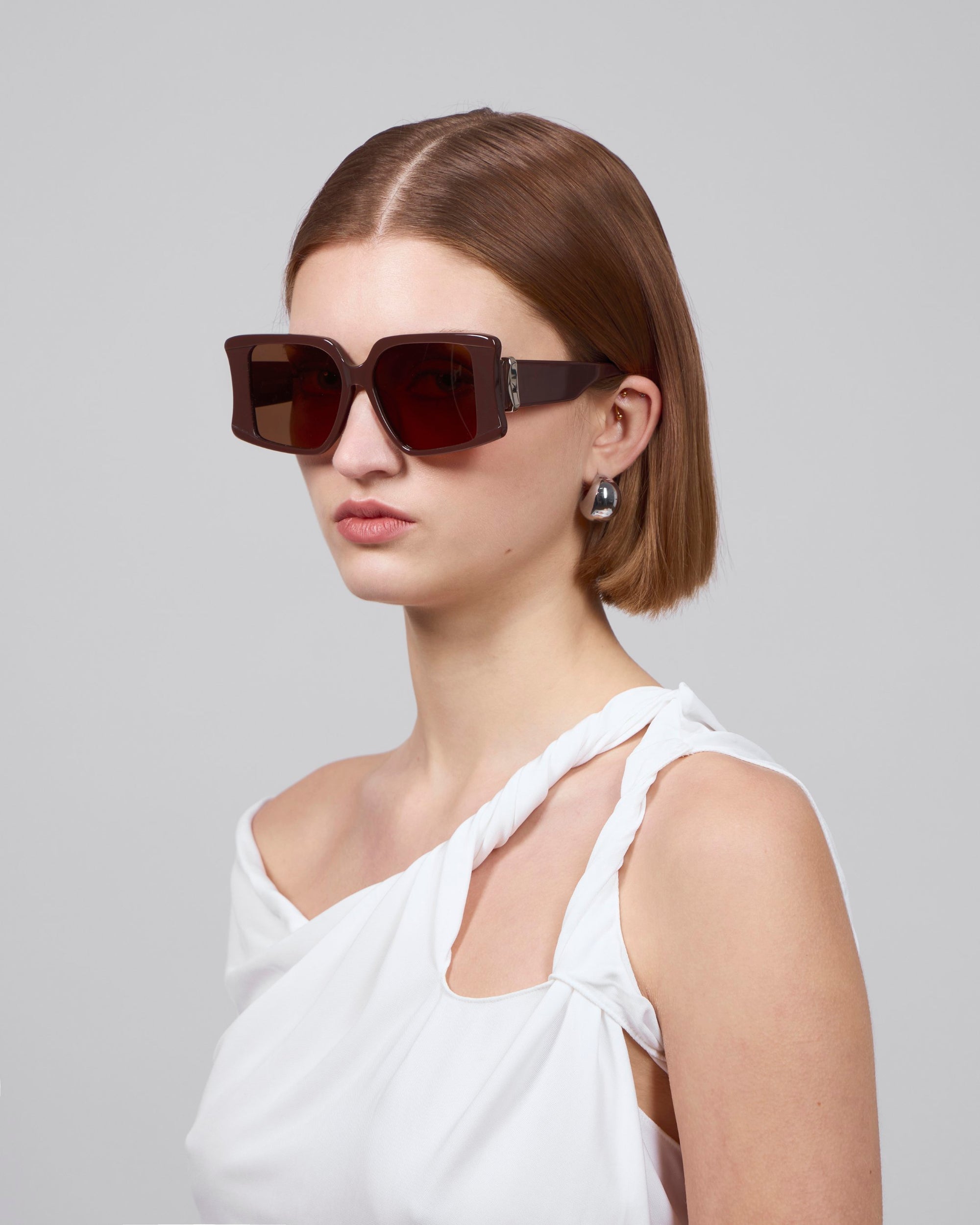 A woman with short brown hair makes a bold statement in her oversized Bloom acetate glasses by For Art's Sake® and white, one-shoulder top. Her pearl earrings add elegance as she poses against a plain background, exuding premium craftsmanship.