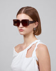 A woman with short brown hair makes a bold statement in her oversized Bloom acetate glasses by For Art's Sake® and white, one-shoulder top. Her pearl earrings add elegance as she poses against a plain background, exuding premium craftsmanship.