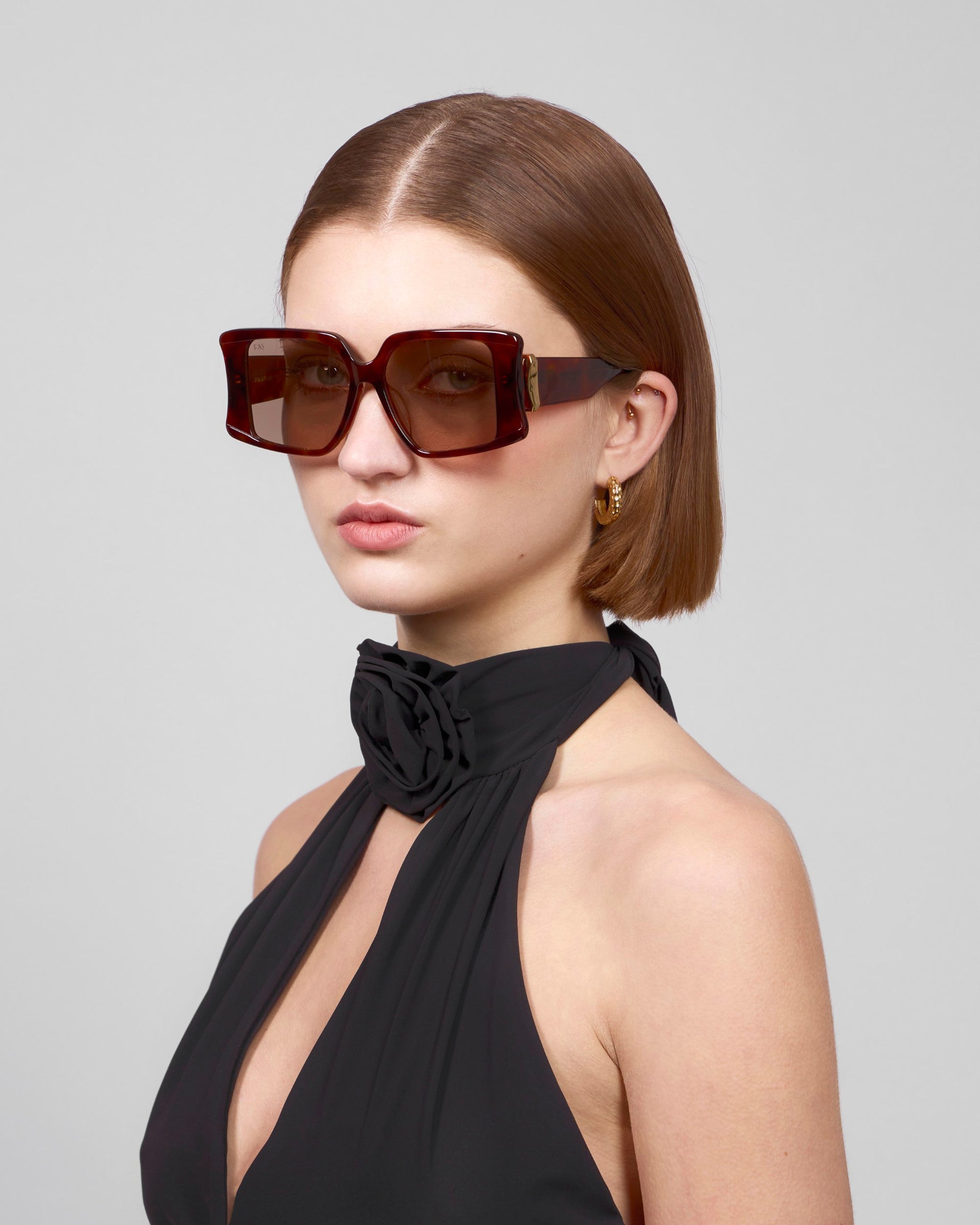 A person with short brown hair wears Bloom's bold statement eyewear from For Art's Sake®—large, square, dark sunglasses—paired with a black halter-neck top featuring a decorative black rose. The backdrop is a plain light gray.