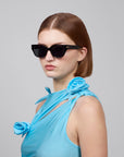 A person with straight brown hair is wearing large, dark cat-eye acetate sunglasses from For Art's Sake® and a sleeveless blue top named Rosie featuring decorative fabric roses on the straps. The background is plain gray.