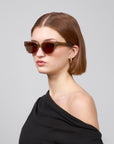 A person with short brown hair wears For Art's Sake® Rosie cat-eye acetate sunglasses and hoop earrings. They're in a black off-the-shoulder top against a plain light gray background, looking to the side.