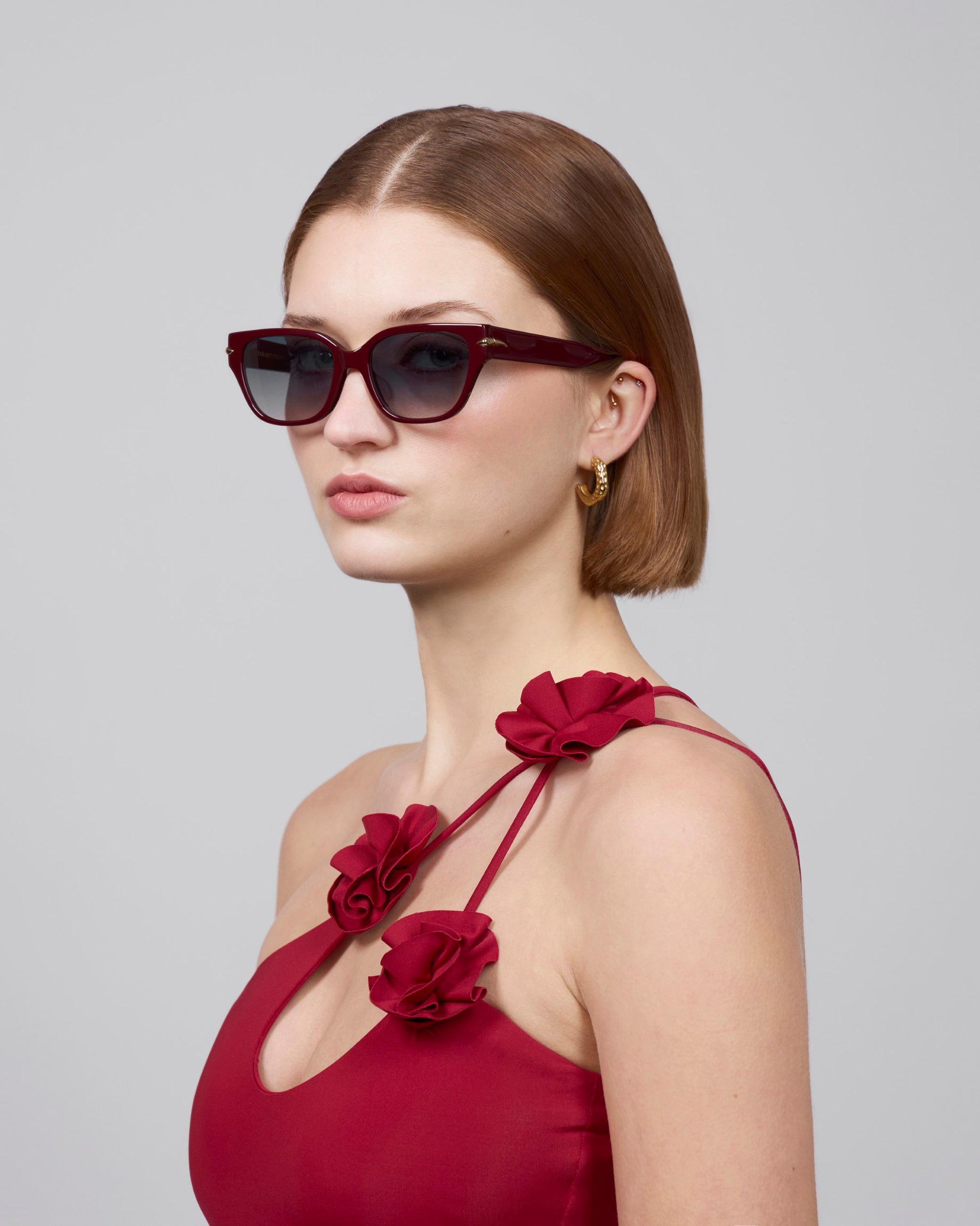 A woman with shoulder-length brown hair poses in Rosie sunglasses from For Art's Sake®, featuring dark cat-eye acetate frames. Her rose-adorned red dress and gold hoop earrings stand out against the plain gray background.