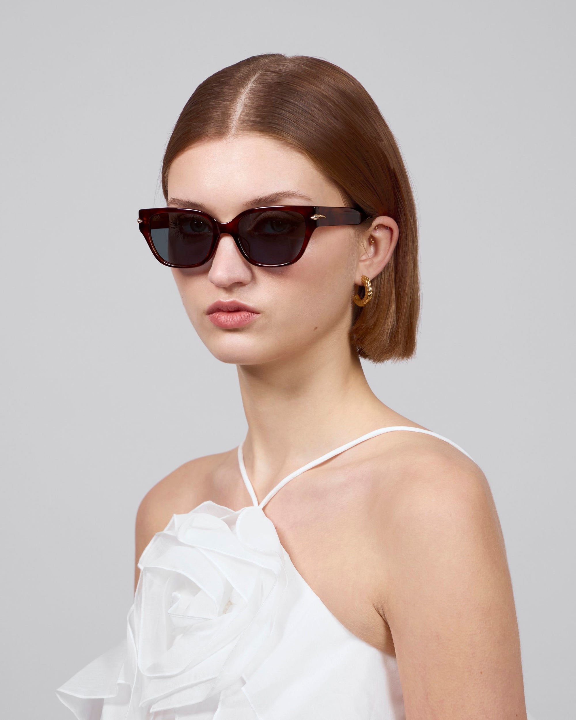 A short-haired individual wears For Art's Sake® Rosie cat-eye acetate sunglasses with a white halter dress featuring a large floral accent. Gold hoop earrings enhance the ensemble, all against a plain gray backdrop, highlighting the chic elegance of the Rose Garden collection.