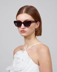 A short-haired individual wears For Art's Sake® Rosie cat-eye acetate sunglasses with a white halter dress featuring a large floral accent. Gold hoop earrings enhance the ensemble, all against a plain gray backdrop, highlighting the chic elegance of the Rose Garden collection.