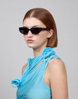 A short-haired person wears For Art's Sake® Lush black cat-eye sunglasses and a floral-decorated light blue top against a plain gray background.