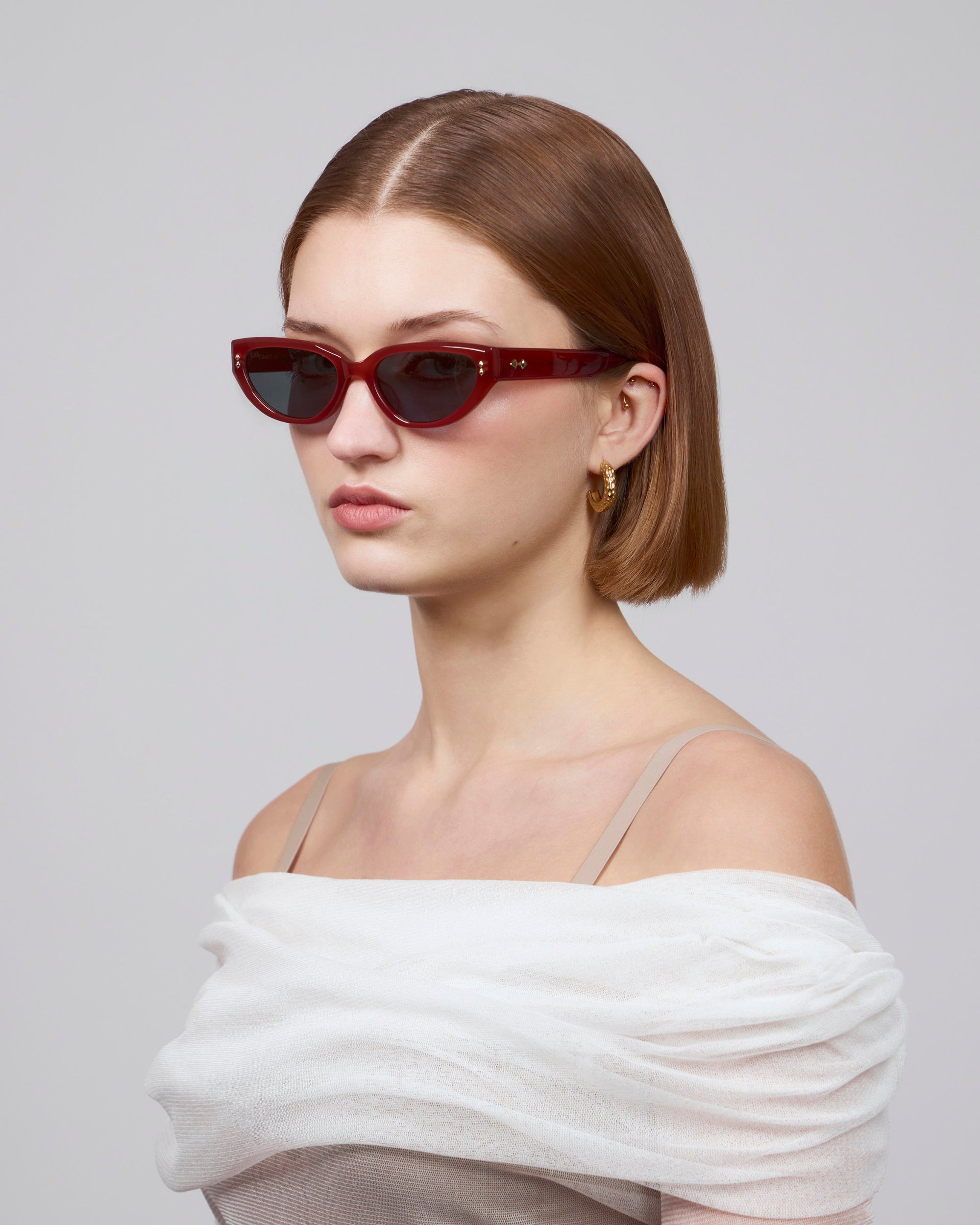 A person with short brown hair sports striking red cat-eye sunglasses, "Lush" by For Art's Sake®, and gold hoop earrings. They wear an off-the-shoulder white top against a plain light gray background.