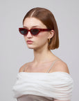 A person with short brown hair sports striking red cat-eye sunglasses, "Lush" by For Art's Sake®, and gold hoop earrings. They wear an off-the-shoulder white top against a plain light gray background.