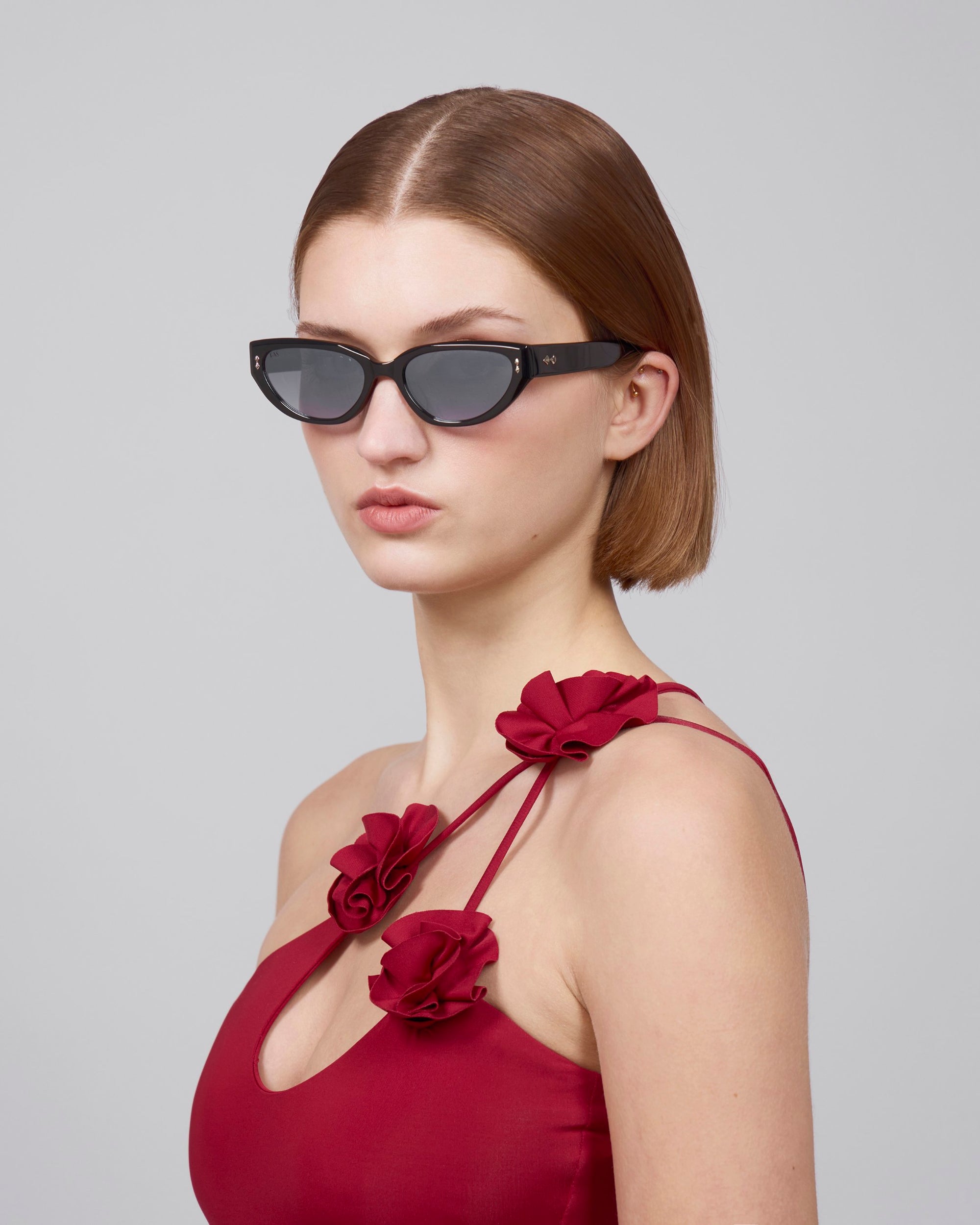 A person with straight brown hair wears For Art's Sake® Lush dark sunglasses and a red dress adorned with large red fabric roses on the straps. The cat-eye frames, crafted from premium materials, beautifully complement the look against a plain, light gray background.