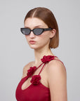 A person with straight brown hair wears For Art's Sake® Lush dark sunglasses and a red dress adorned with large red fabric roses on the straps. The cat-eye frames, crafted from premium materials, beautifully complement the look against a plain, light gray background.