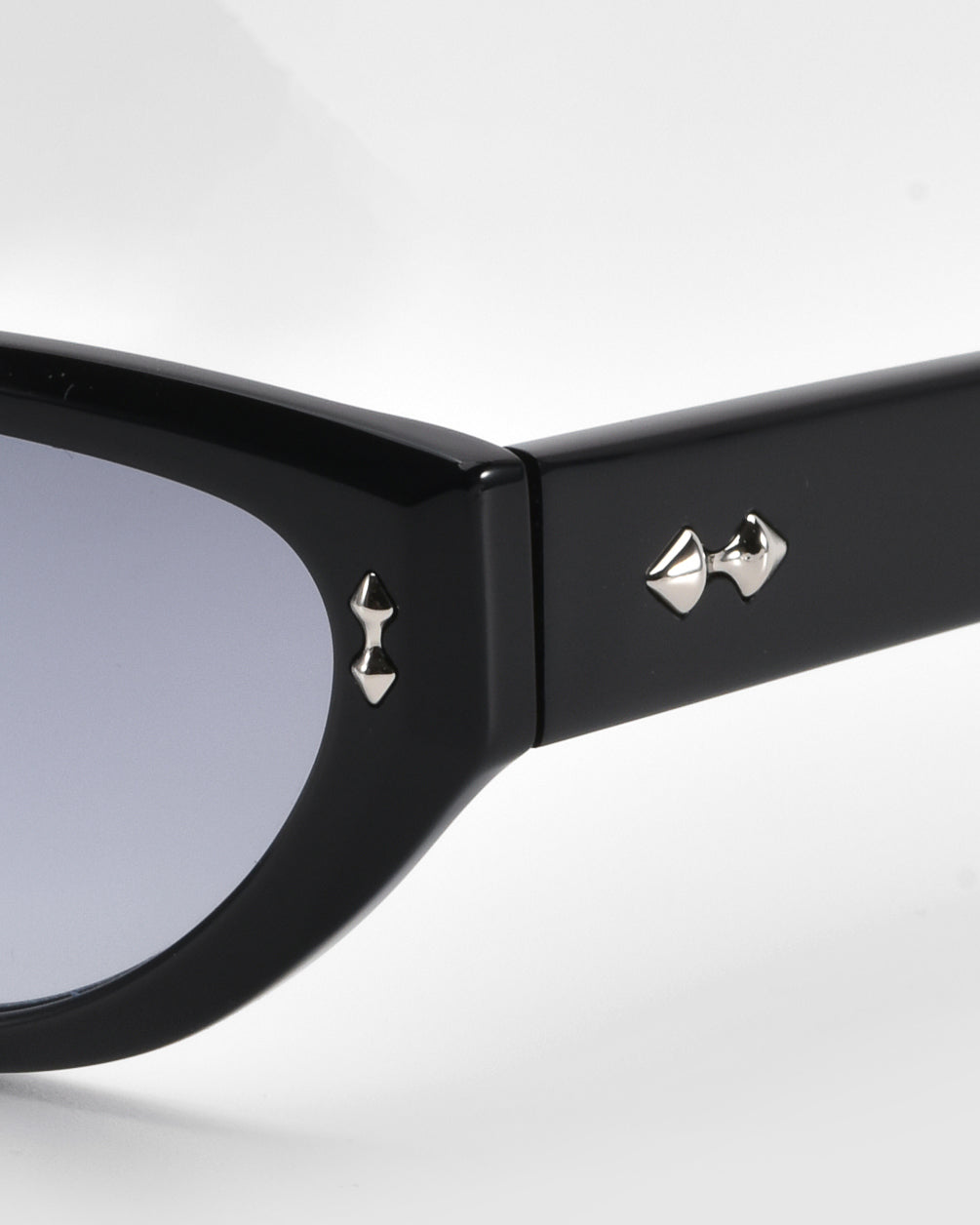 Close-up of the Lush sunglasses by For Art's Sake® features black cat-eye style with a glossy finish, flaunting small silver pyramid studs on the temples near the hinge. Crafted from premium materials, these sunglasses have dark-tinted lenses for a sleek and modern look.