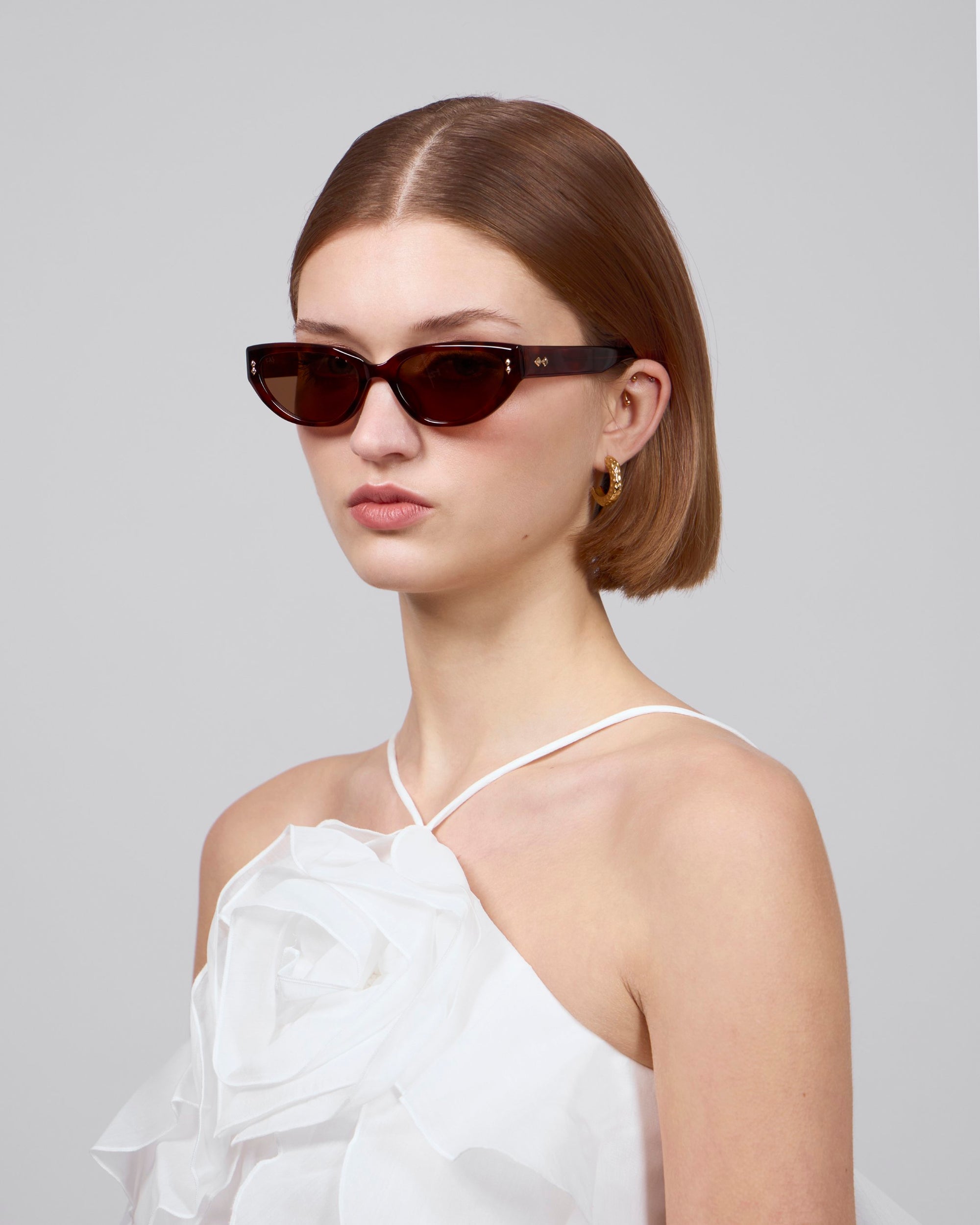 A person with short, light brown hair wears Lush sunglasses by For Art&#39;s Sake® and a white halter dress with a large floral design on the bodice. The background is plain gray.