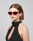 A person with short brown hair is wearing red-tinted Lush cat-eye sunglasses by For Art's Sake®, gold hoop earrings, and a black halter dress featuring a flower detail. They are posed against a plain gray background.