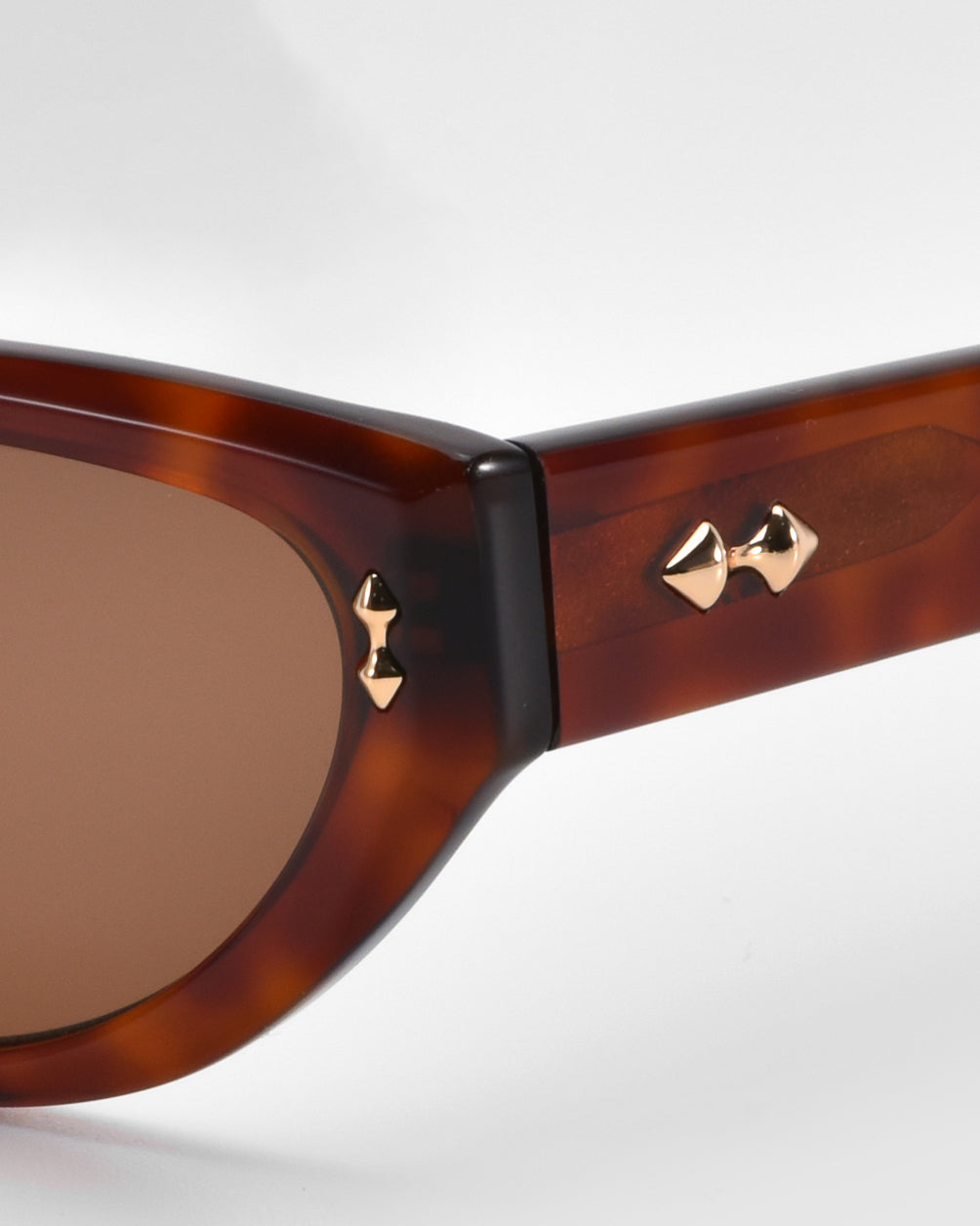 Close-up of For Art's Sake® Lush sunglasses in a brown cat-eye style, with brass bow tie-shaped studs near the hinge and dark-tinted lenses. The premium tortoiseshell-patterned frame stands out against a minimalist white background.