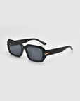 The Fleur sunglasses by For Art's Sake® feature stylish black rectangular acetate frames with dark lenses. Thick arms boast elegant gold accents near the hinges, all set against a serene plain light gray background reminiscent of a Rose Garden on a sunny afternoon.