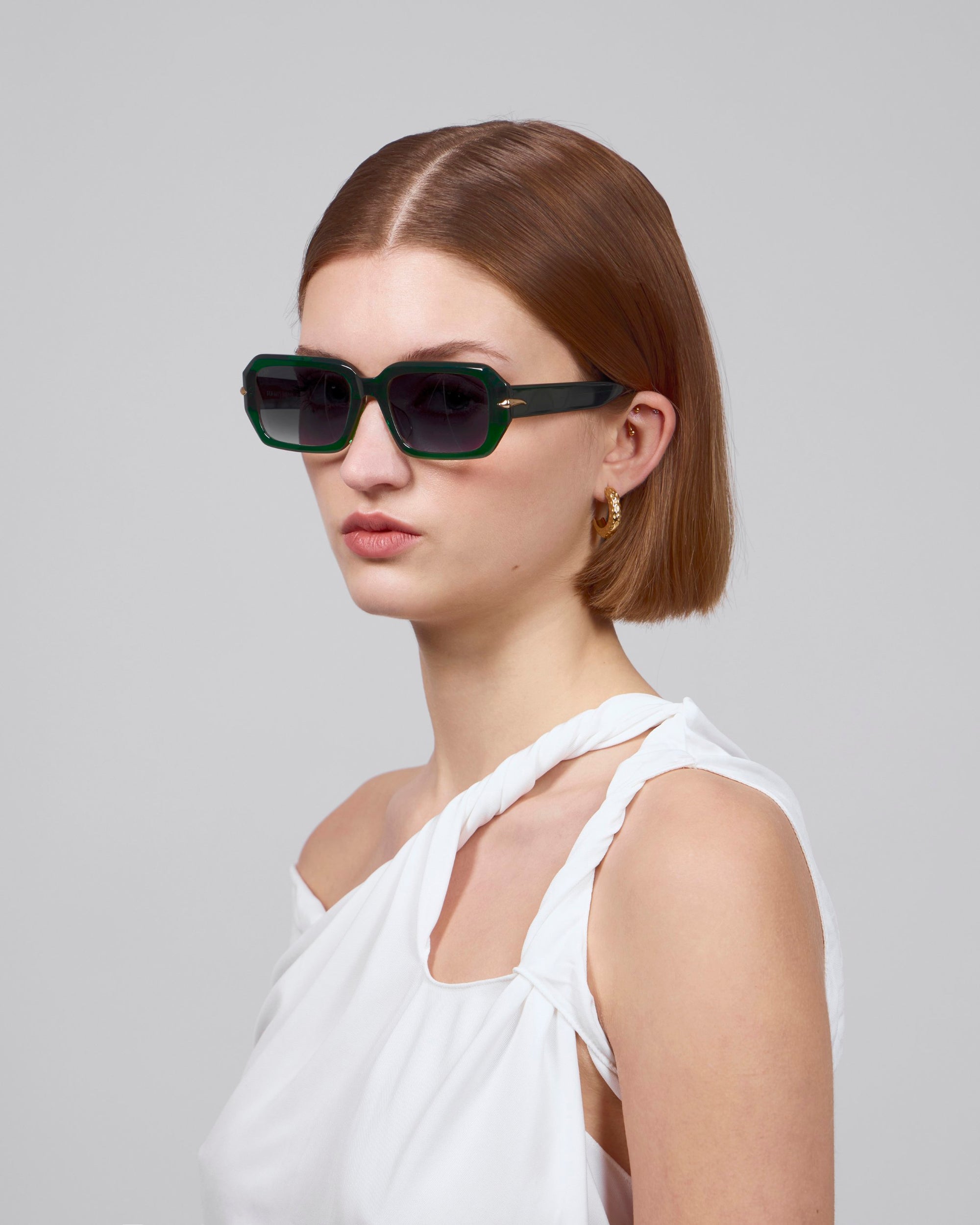 A short-haired person wears Fleur green sunglasses by For Art's Sake®, pairing them with a chic white asymmetrical top against a plain light gray background.