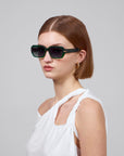 A short-haired person wears Fleur green sunglasses by For Art's Sake®, pairing them with a chic white asymmetrical top against a plain light gray background.