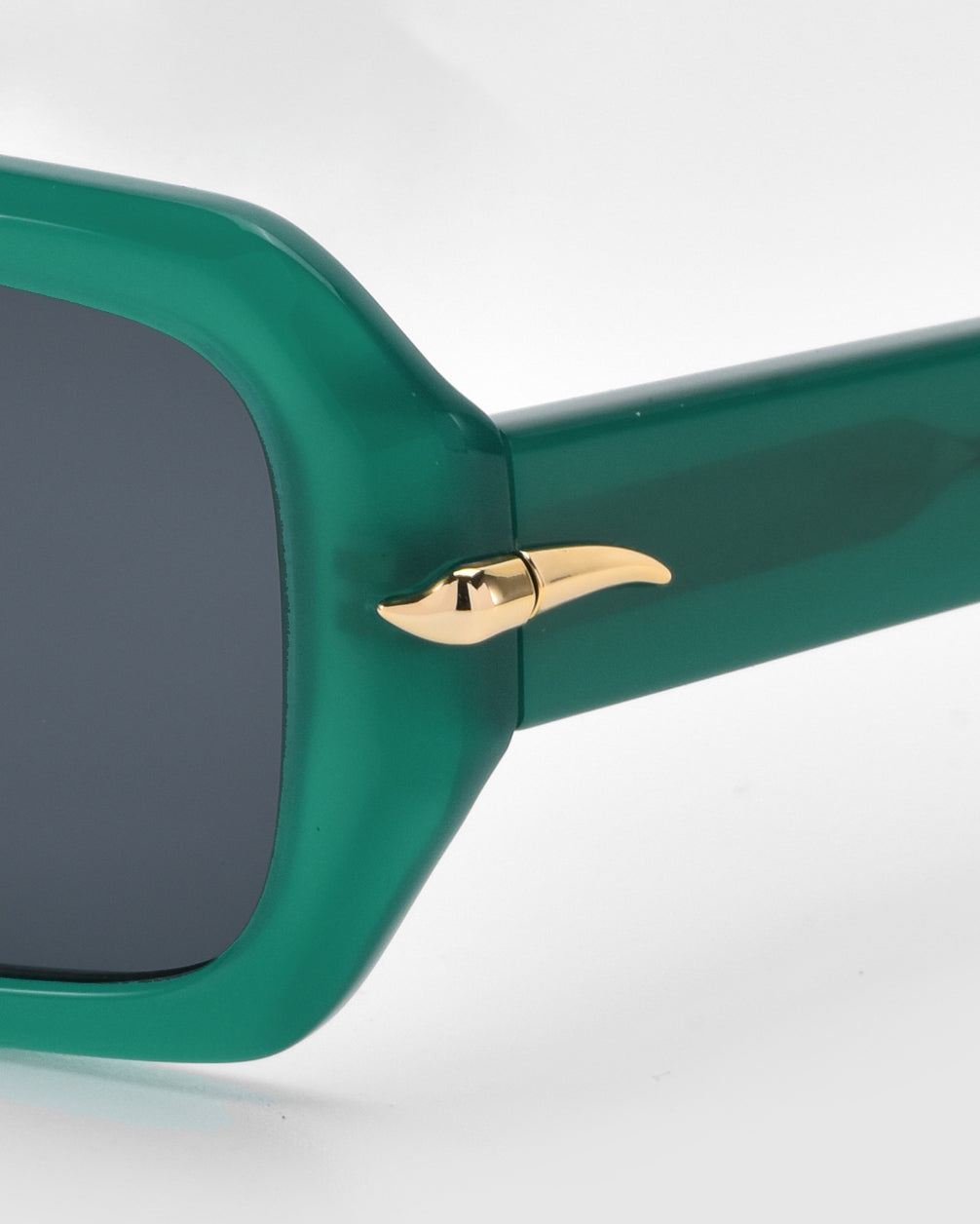 A close-up of the For Art's Sake® Fleur sunglasses shows a minimalist green frame with tinted lenses. The temple hinge features a gold metallic accent resembling a horn or curved spike, set against a white background.