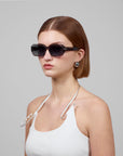 A person with chin-length brown hair and pearl earrings showcases a minimalist Fleur design in profile. Dark sunglasses by For Art's Sake® enhance their look, paired with a white sleeveless top and cord necklace, set against a gray background.