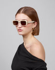 A person with straight, shoulder-length hair wearing sleek rectangular Fleur sunglasses by For Art's Sake® and a black off-the-shoulder top, gazing to the side against a minimalist background.